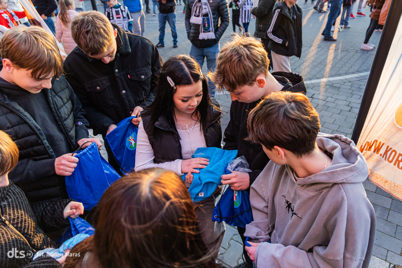 Group of teenagers gathering blue event bags from a distribution table in a sunny plaza.