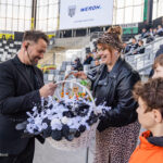 Man with headset hands a large white flower basket to a smiling woman and three children in a stadium.