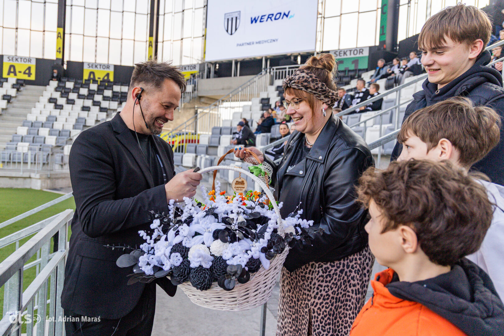 Man with headset hands a large white flower basket to a smiling woman and three children in a stadium.