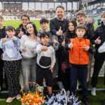 Group of kids and adults on a soccer field posing for a photo, giving thumbs up, with baskets of orange and white flowers.