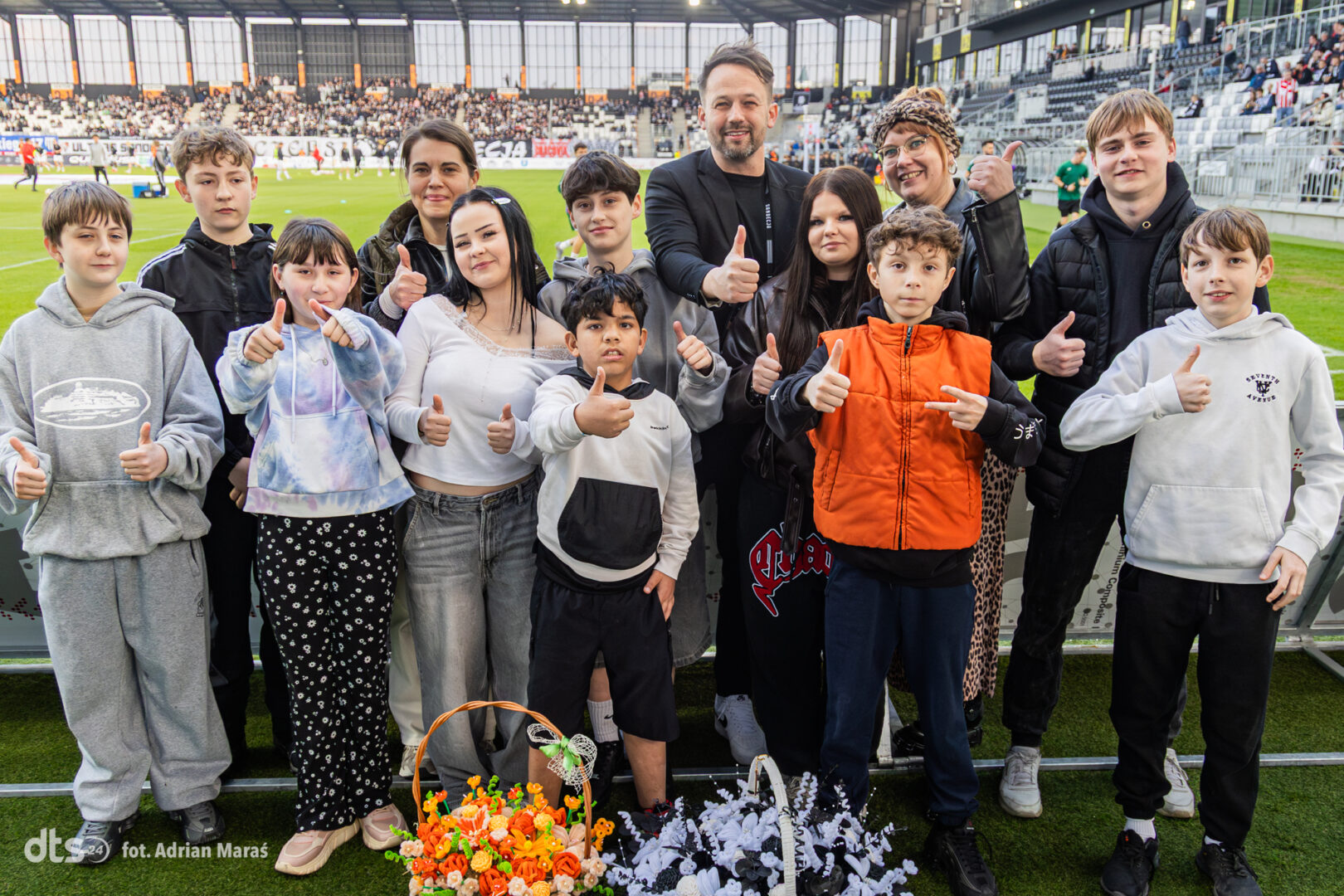 Group of kids and adults on a soccer field posing for a photo, giving thumbs up, with baskets of orange and white flowers.