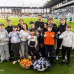 Group of kids and adults posing with thumbs up on a soccer field, flower baskets in front, stadium stands behind.