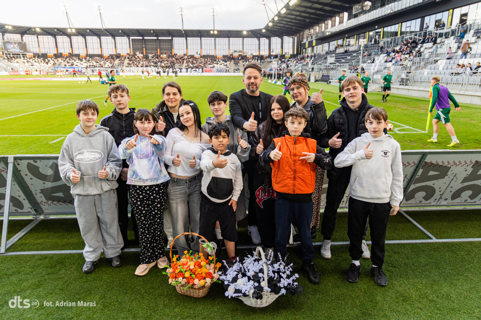 Group of kids and adults posing with thumbs up on a soccer field, flower baskets in front, stadium stands behind.