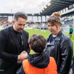 Man in a black blazer shakes hands with a child wearing an orange vest on a football pitch, with a woman in a leather jacket and a teen nearby in the background stands.