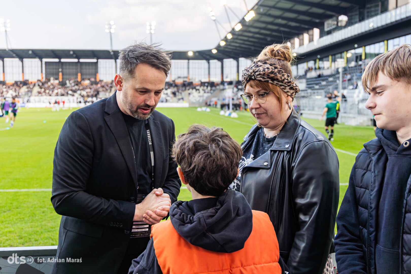 Man in a black blazer shakes hands with a child wearing an orange vest on a football pitch, with a woman in a leather jacket and a teen nearby in the background stands.
