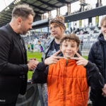 Group of three youths and a man near the stadium sideline, with the boy in an orange jacket flashing peace signs at the camera.