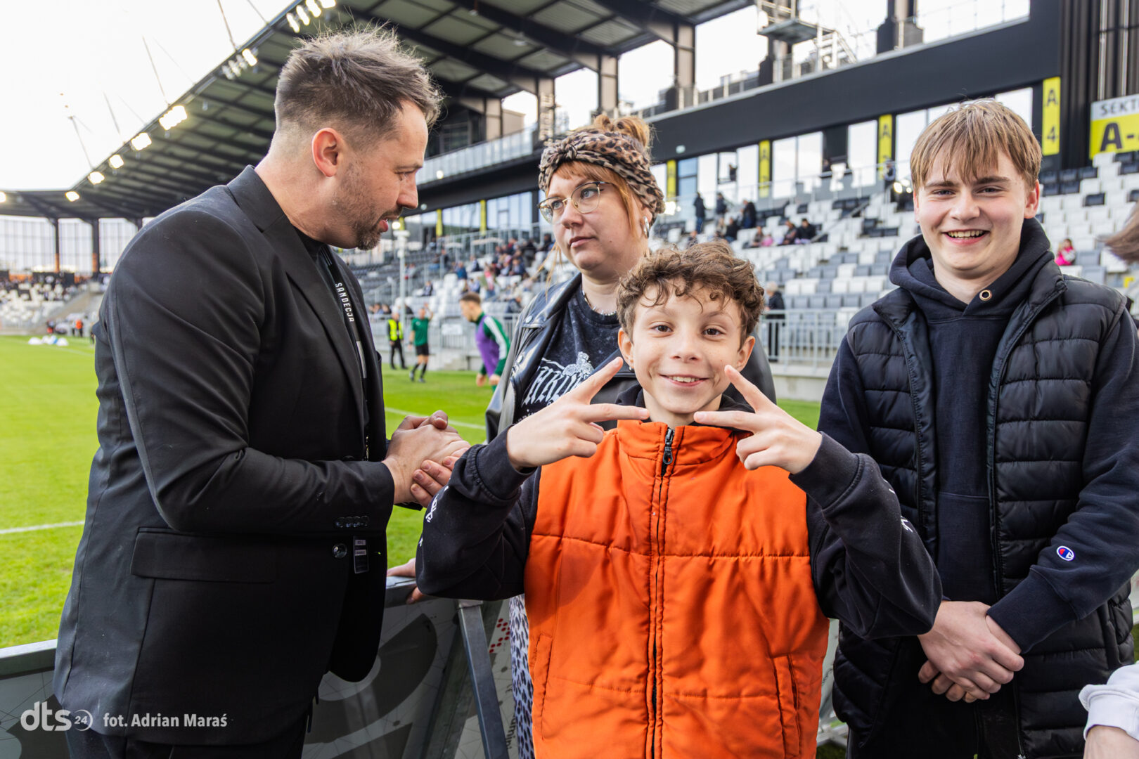 Group of three youths and a man near the stadium sideline, with the boy in an orange jacket flashing peace signs at the camera.