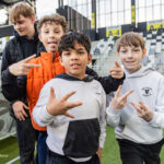 Four boys posing together at a soccer stadium, making playful hand signs and smiling at the camera as they stand on the sideline near the field.