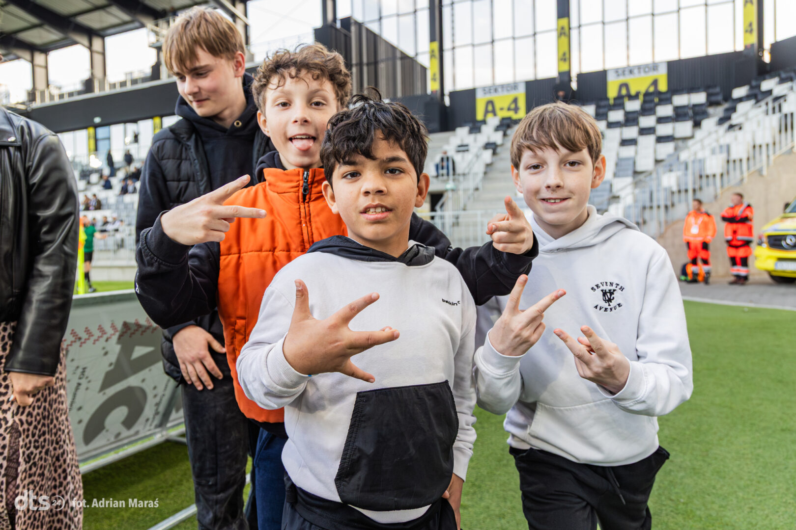 Four boys posing together at a soccer stadium, making playful hand signs and smiling at the camera as they stand on the sideline near the field.
