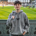 Youth posing in a gray hoodie at a soccer stadium, fists raised in a casual pose with a green field behind him