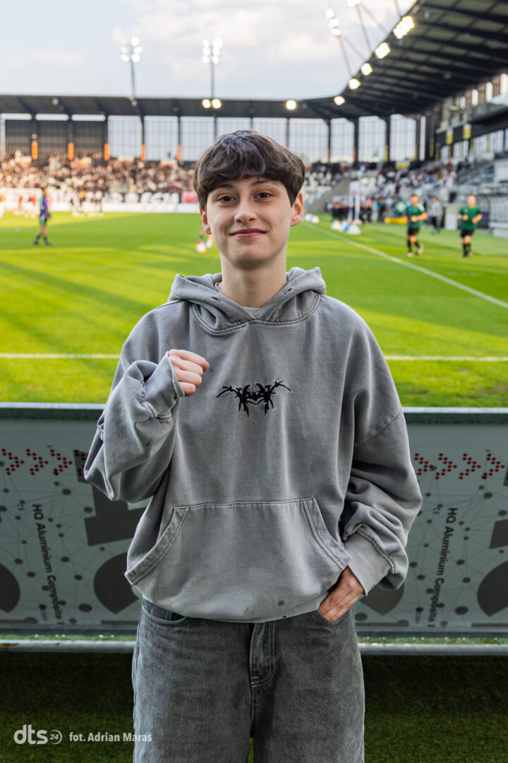 Youth posing in a gray hoodie at a soccer stadium, fists raised in a casual pose with a green field behind him
