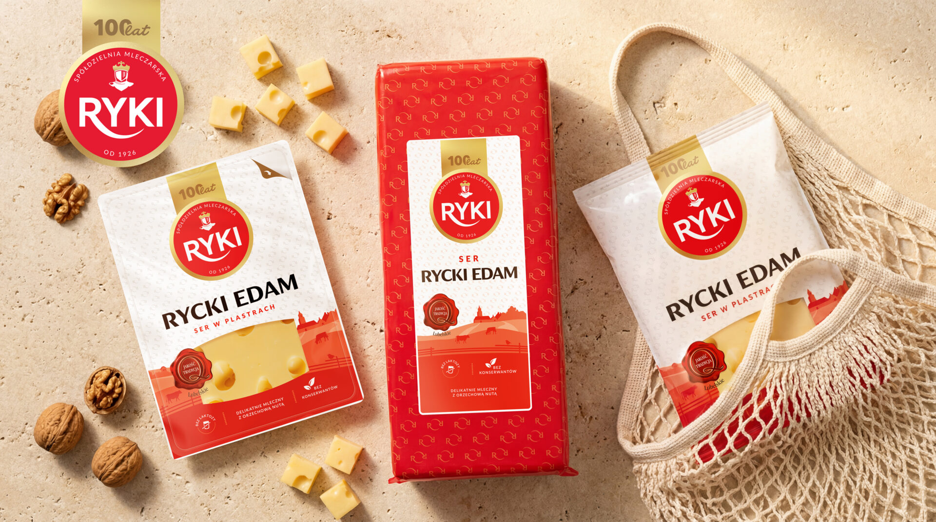 Assortment of Rycki Edam cheese packaging on a beige surface, featuring a red gift box, white cheese pouches, scattered cheese cubes, walnuts, and a beige tote bag with the Rycki logo visible.