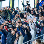 Crowd of sports fans in a stadium stands, many with arms raised in cheers, wearing jackets and scarves.
