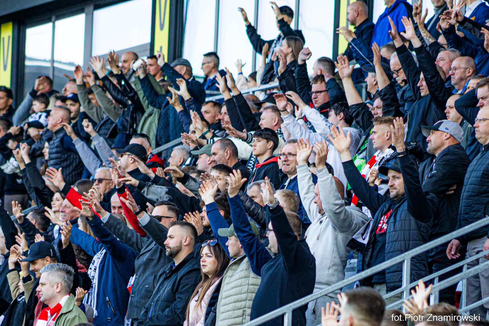 Crowd of sports fans in a stadium stands, many with arms raised in cheers, wearing jackets and scarves.