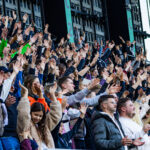 Crowd of spectators in a stadium with many hands raised in the air, cheering for a live event.