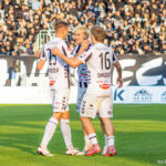 Soccer players in white jerseys hug and celebrate on a green field as a cheering crowd looks on from the stands.