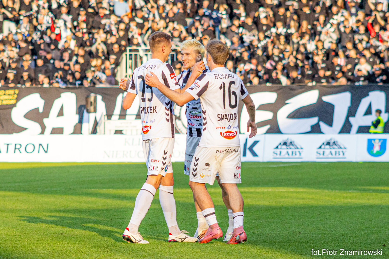 Soccer players in white jerseys hug and celebrate on a green field as a cheering crowd looks on from the stands.