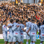Soccer players in white uniforms celebrate on the field as a packed crowd cheers with scarves in the stands behind them.