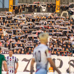 Tall stadium stands filled with fans holding black-and-white scarves aloft, creating a sea of supporters behind the goal.