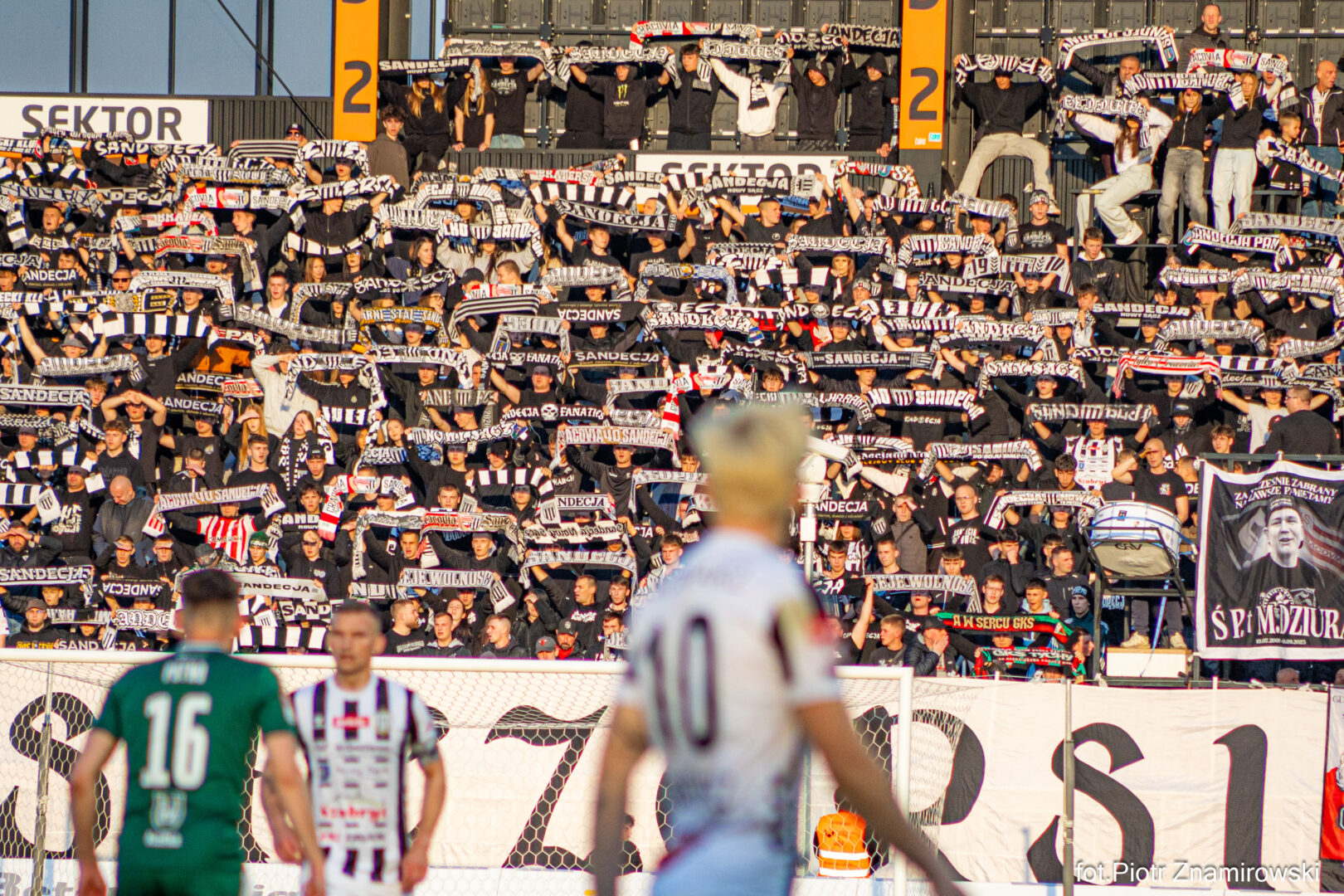 Tall stadium stands filled with fans holding black-and-white scarves aloft, creating a sea of supporters behind the goal.