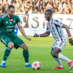 Two soccer players contesting the ball on a green field, one in green kit and the other in white with black stripes, as they move toward a small red-and-white ball.