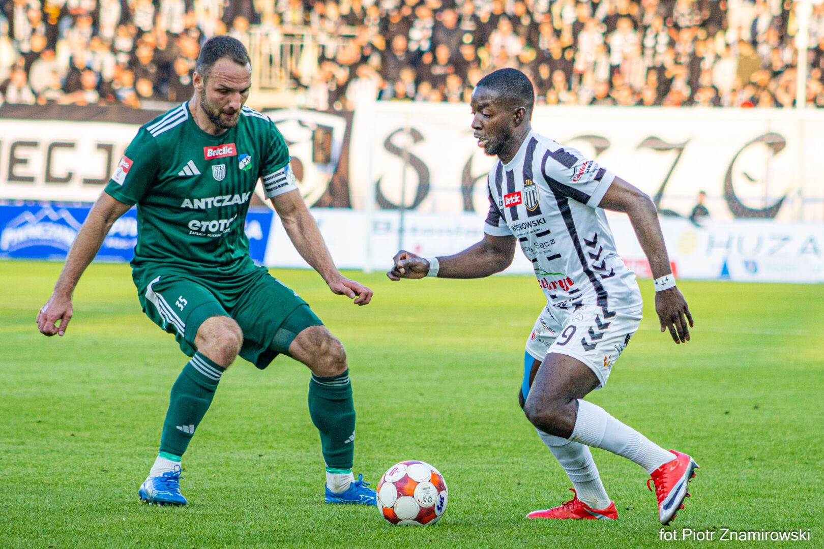 Two soccer players contesting the ball on a green field, one in green kit and the other in white with black stripes, as they move toward a small red-and-white ball.