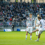 Two players in white-and-black striped uniforms contest the ball on a green soccer field, with a packed stadium in the background.