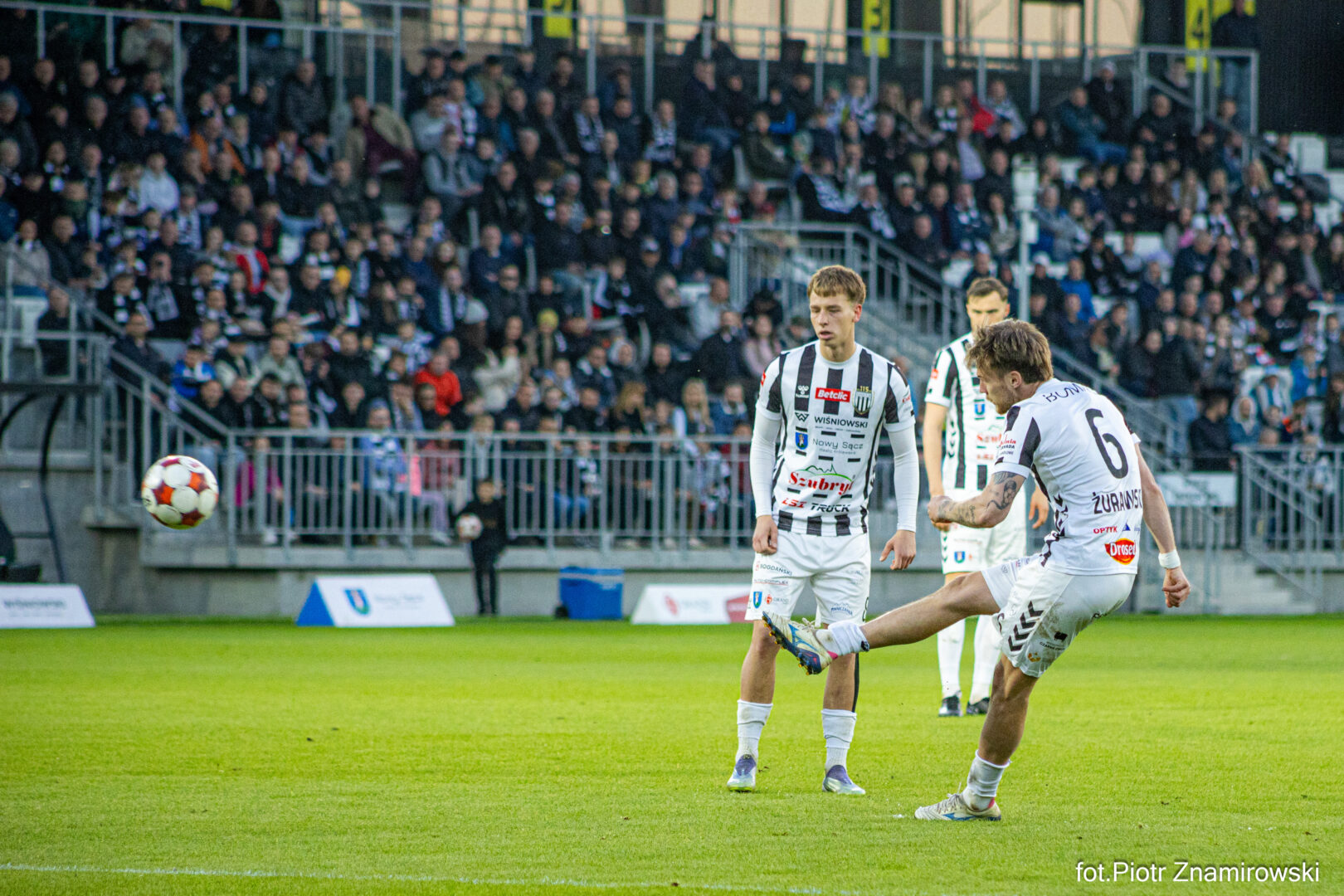 Two players in white-and-black striped uniforms contest the ball on a green soccer field, with a packed stadium in the background.