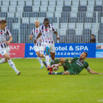 Soccer scene with a green-clad goalie sliding to tackle a ball while white-and-purple striped players approach.]