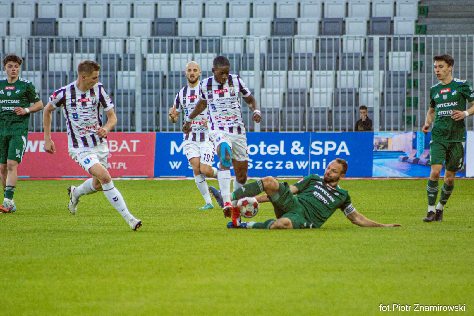 Soccer scene with a green-clad goalie sliding to tackle a ball while white-and-purple striped players approach.]
