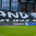 Giant black and white banner held by fans across the stands, featuring bold white letters and a skull emblem on the right, with a green pitch in the foreground.