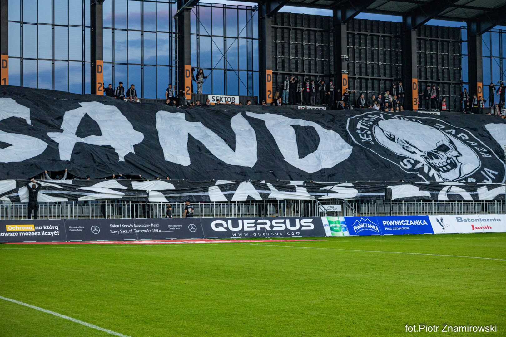 Giant black and white banner held by fans across the stands, featuring bold white letters and a skull emblem on the right, with a green pitch in the foreground.