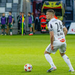 Soccer player in a white kit with number 24 controls a ball on a green pitch, with medical staff and substitutes on the sideline in the background.
