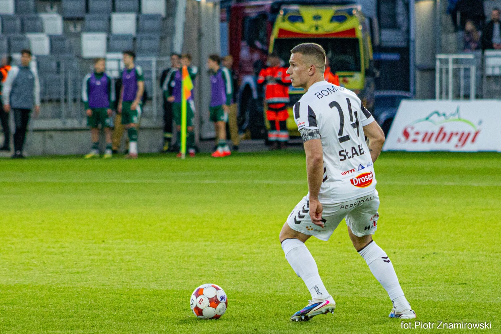 Soccer player in a white kit with number 24 controls a ball on a green pitch, with medical staff and substitutes on the sideline in the background.