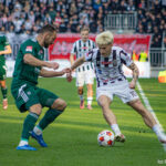 Two soccer players battle for the ball on a sunny pitch while a crowd watches from the stands behind them.