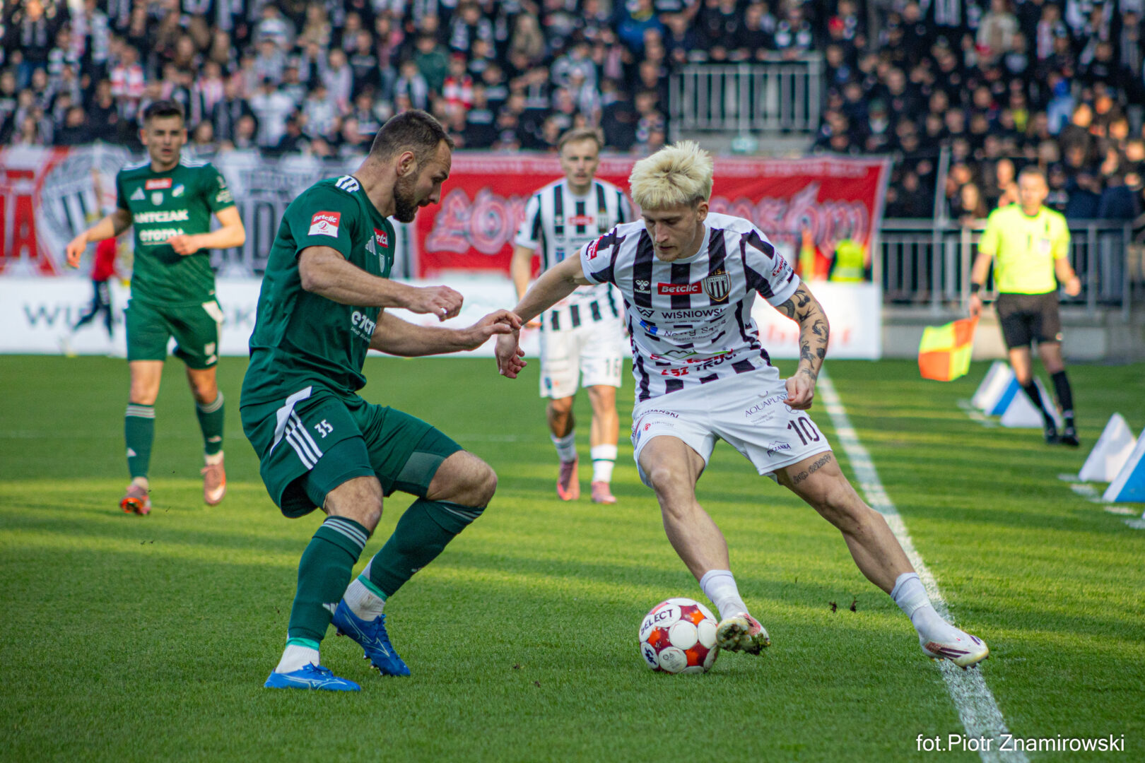 Two soccer players battle for the ball on a sunny pitch while a crowd watches from the stands behind them.