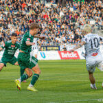 Football match moment: white-jersey player (number 10) dribbling toward the goal as two green-jersey players close in, with a crowded stadium in the background