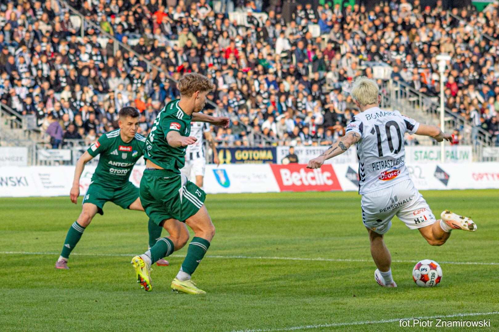 Football match moment: white-jersey player (number 10) dribbling toward the goal as two green-jersey players close in, with a crowded stadium in the background