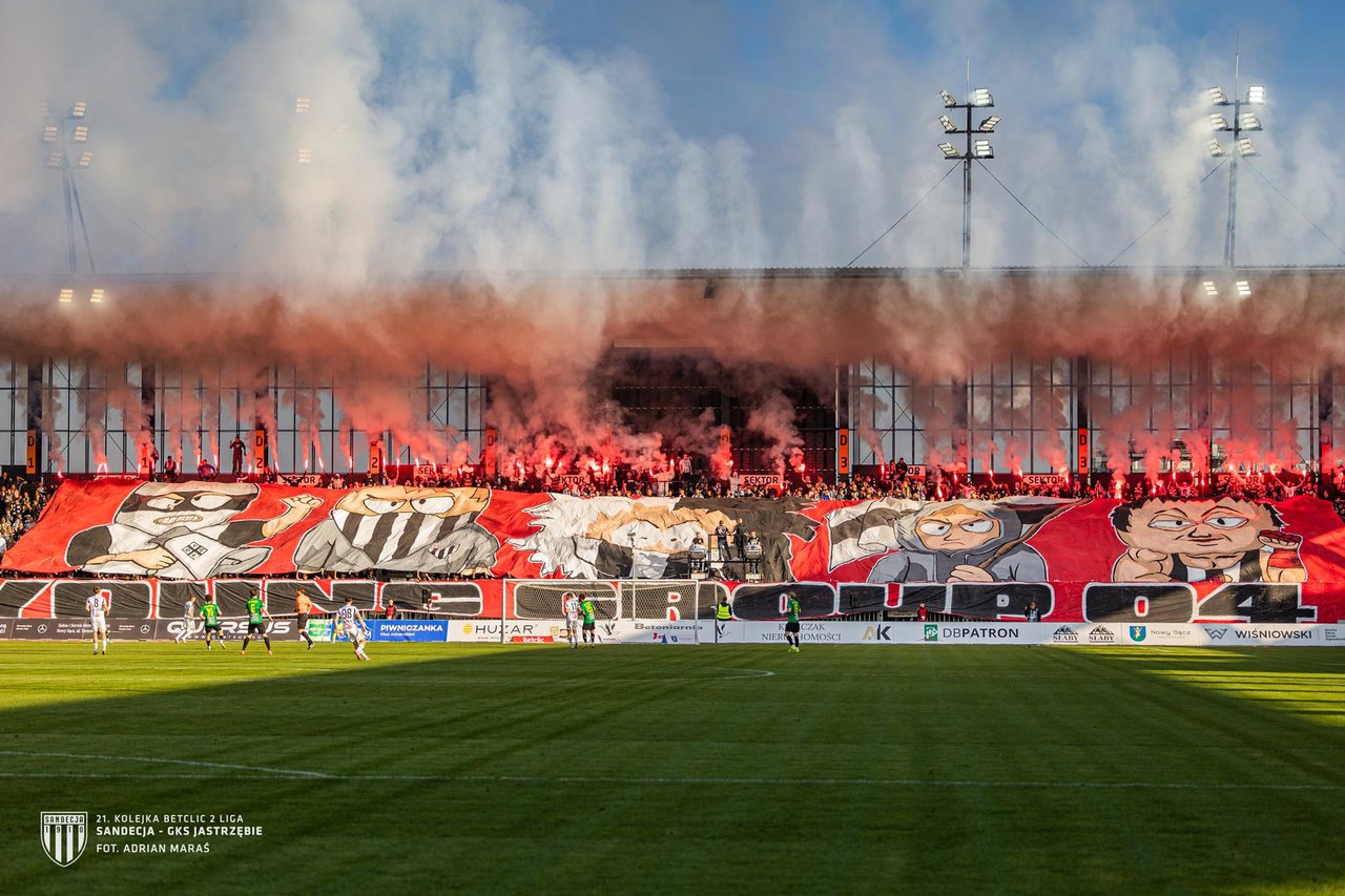 Crowd of red-clad football fans in a stadium with a giant illustrated banner across the stands and red smoke rising above them.