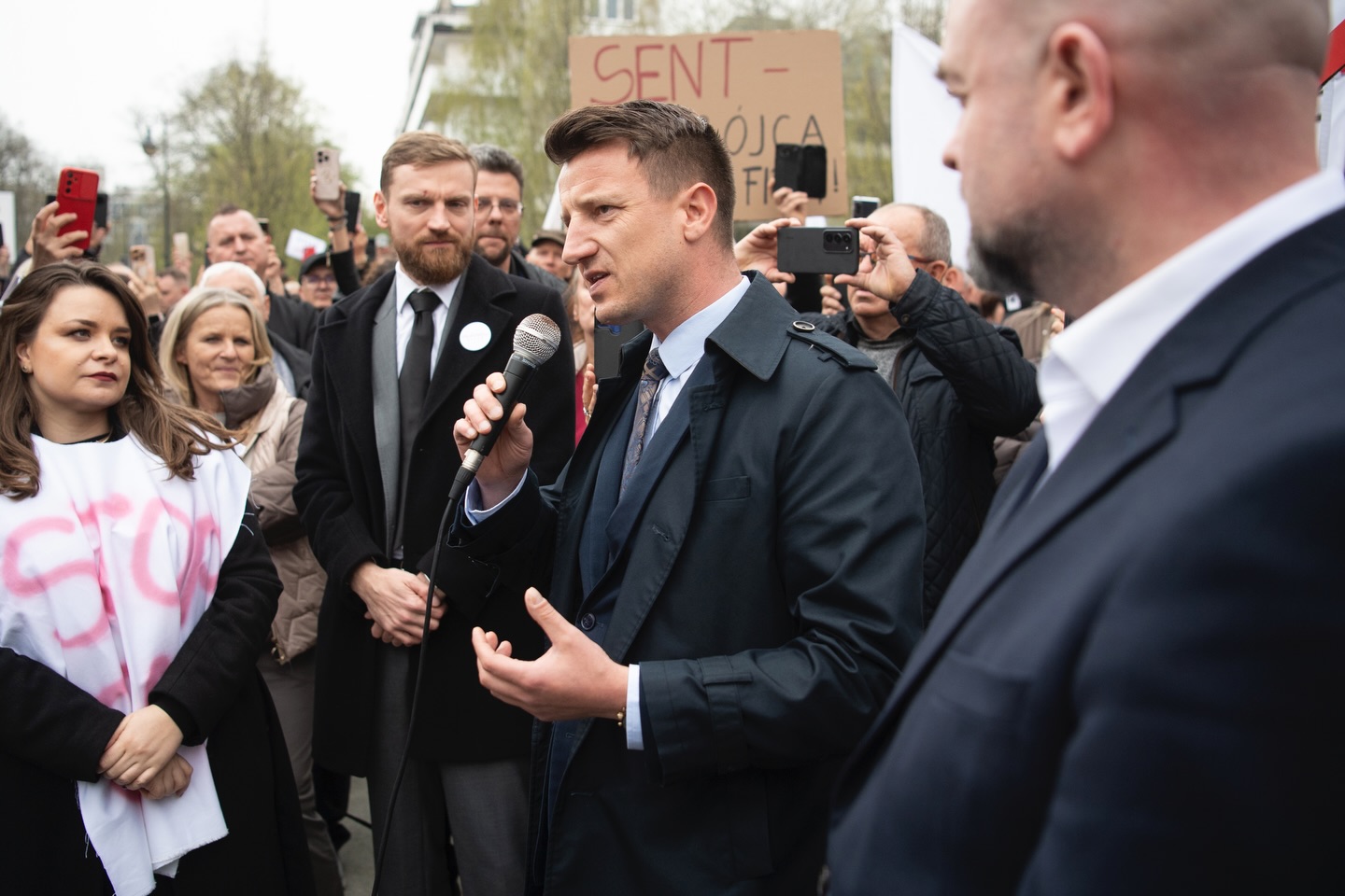 Man in a dark coat speaks into a handheld microphone at a street protest, surrounded by a crowd recording with phones and holding signs.