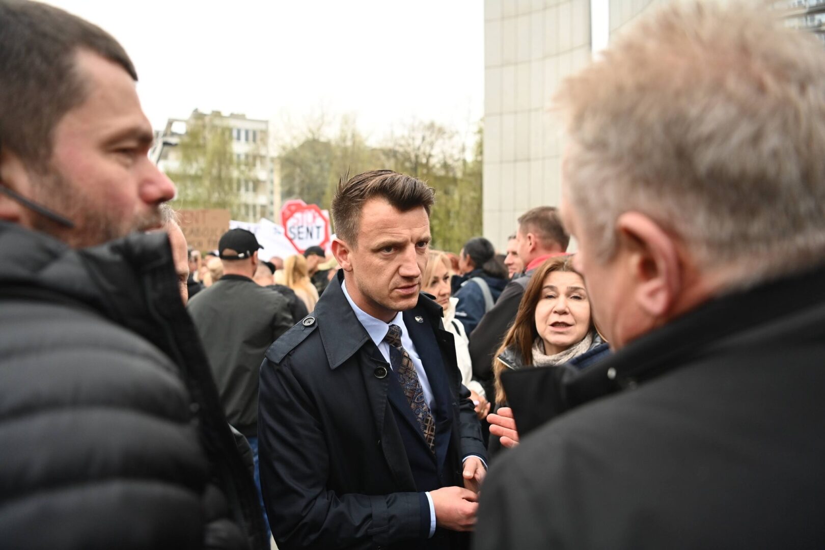 Man in a dark coat and tie talking to a crowd at a street protest with bystanders and signs in the background.