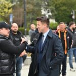 Two men shake hands in the foreground: one wearing a black puffer vest and cap, the other in a dark suit and coat, with a group watching behind them.