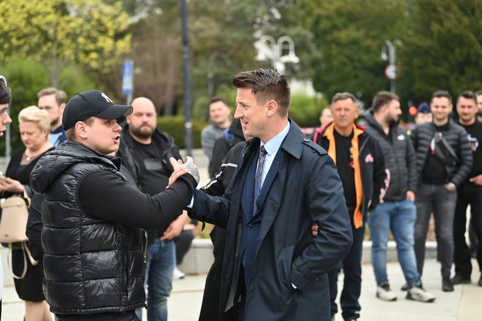 Two men shake hands in the foreground: one wearing a black puffer vest and cap, the other in a dark suit and coat, with a group watching behind them.
