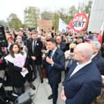 Man in a navy suit speaks into a microphone at a crowded protest rally, with signs visible in the background (e.g., STOP SENT).