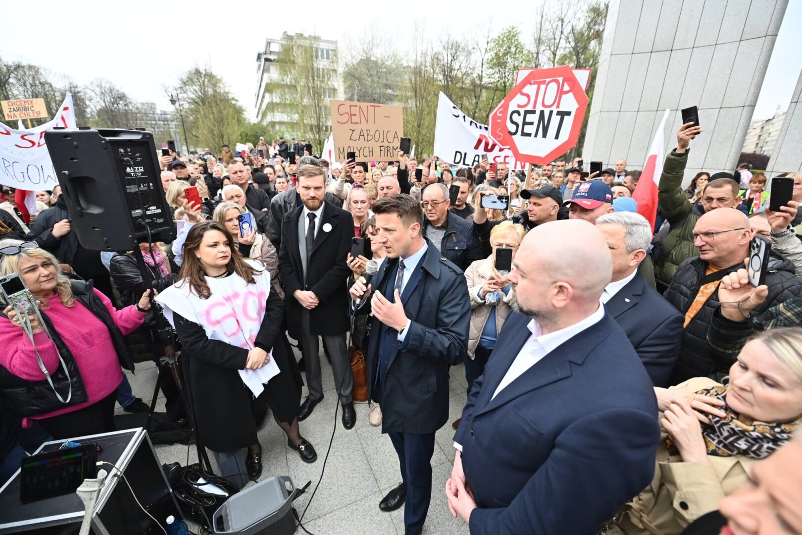 Man in a navy suit speaks into a microphone at a crowded protest rally, with signs visible in the background (e.g., STOP SENT).