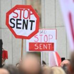 Red-and-white octagonal stop sign prop reading 'STOP SENT' raised above a crowd at a protest event.