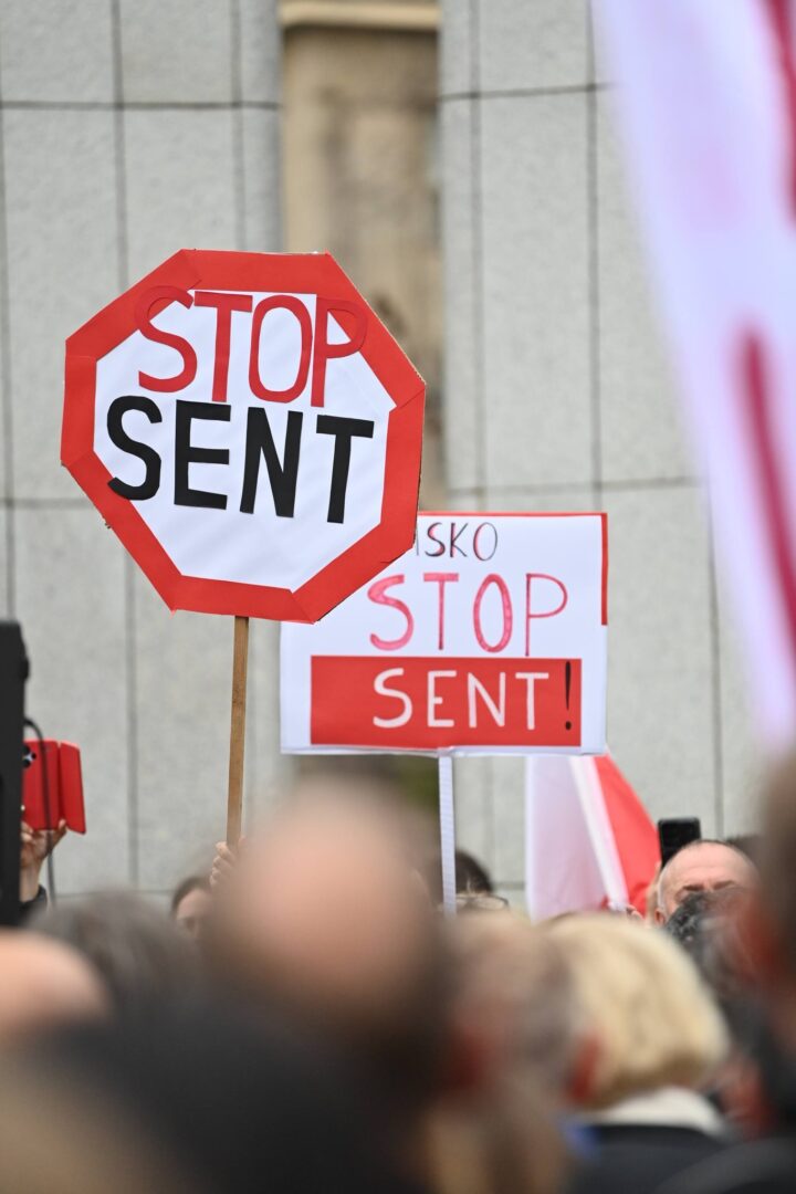Red-and-white octagonal stop sign prop reading 'STOP SENT' raised above a crowd at a protest event.