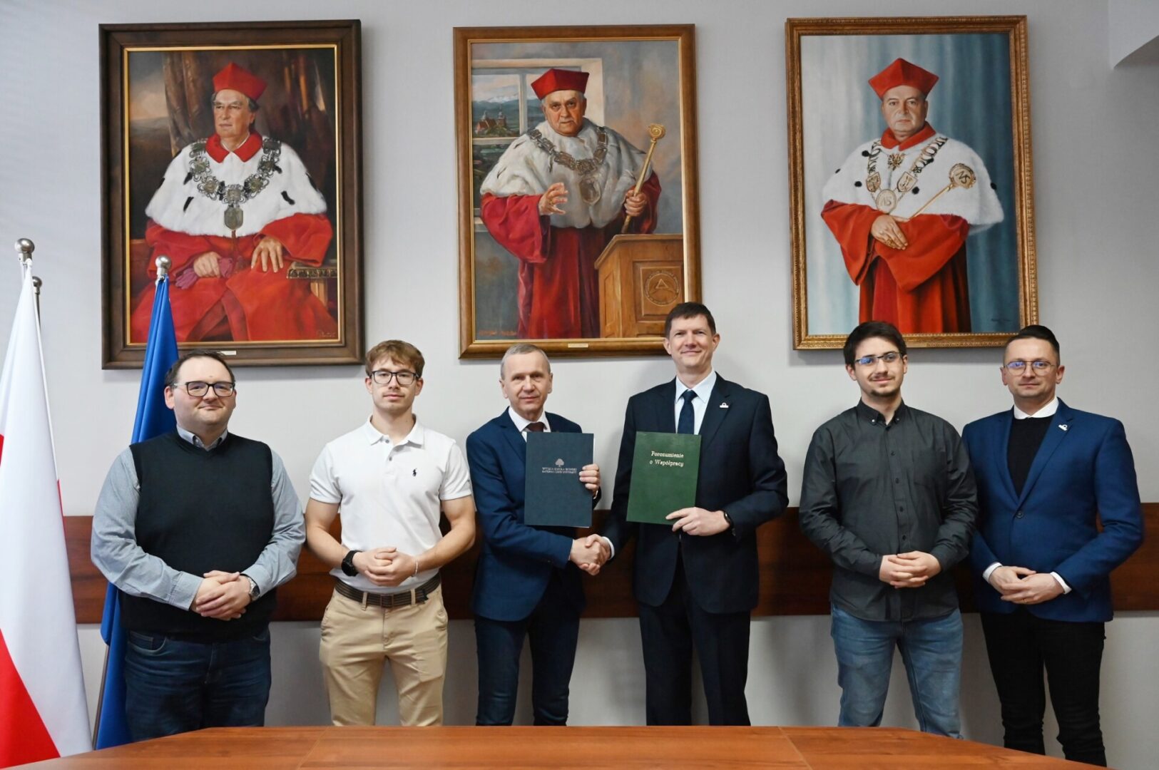 Six men in business attire pose in a formal room; two shake hands while holding documents, with flags and three framed portraits behind them.