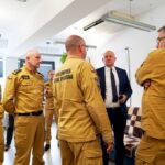 Group of Polish State Fire Service firefighters in tan uniforms listening to a man in a suit indoors.