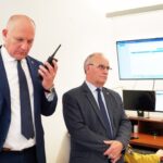 Two men in suits in a room with computer monitors; one holds a handheld radio while the other watches attentively at a desk setup.